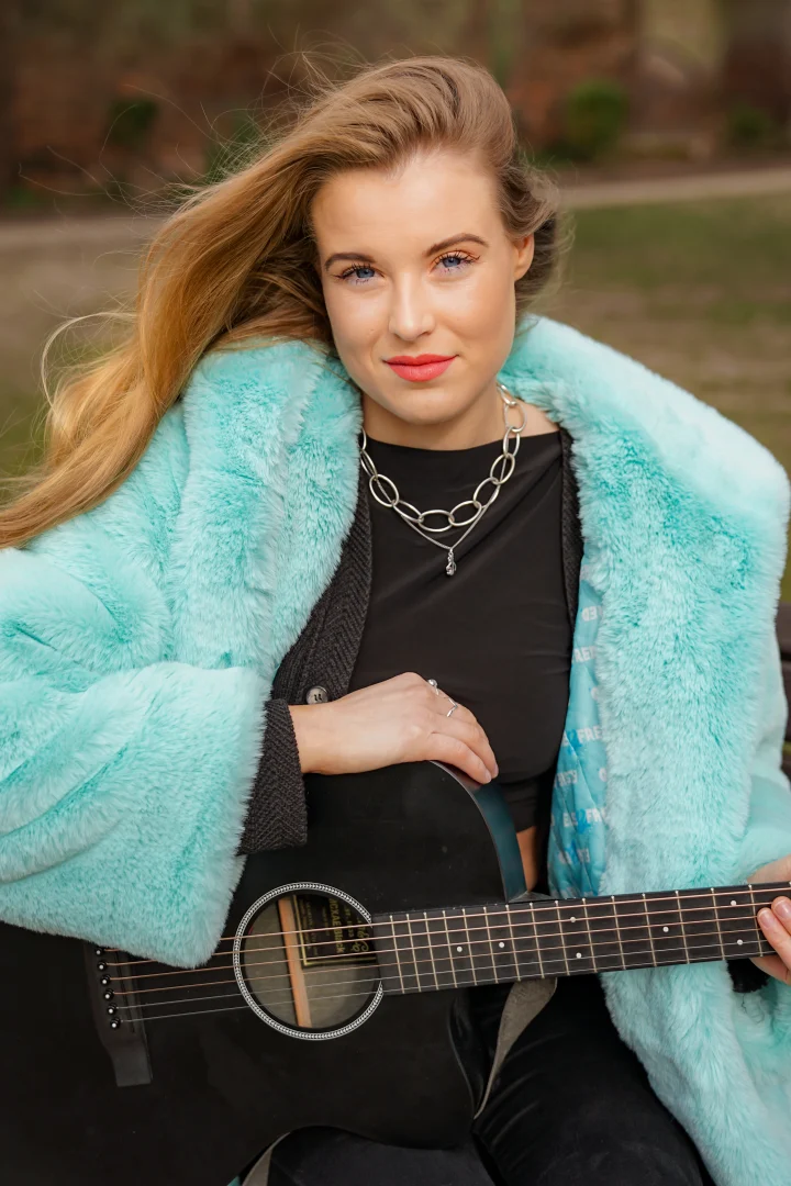 Grace George smiling at the camera in an aqua faux-fur coat and chain necklace, holding a black guitar outdoors.