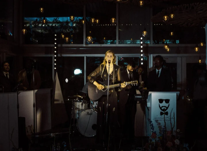Grace George singing with a guitar in a dim venue, band members in suits and a drum kit behind her.