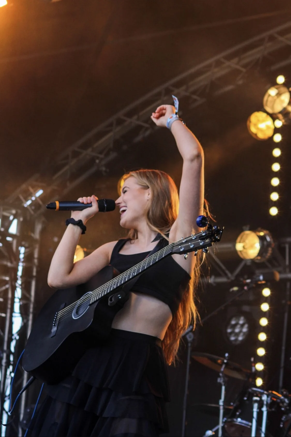 Grace George laughing on a festival stage with one arm raised, holding a microphone and black guitar, stage lights behind.
