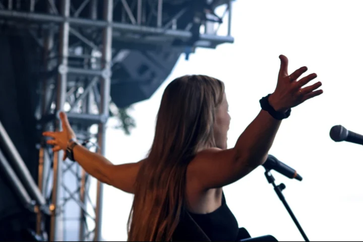 Grace George seen from behind on an outdoor stage, arms spread wide toward a bright sky, scaffolding and microphone nearby.