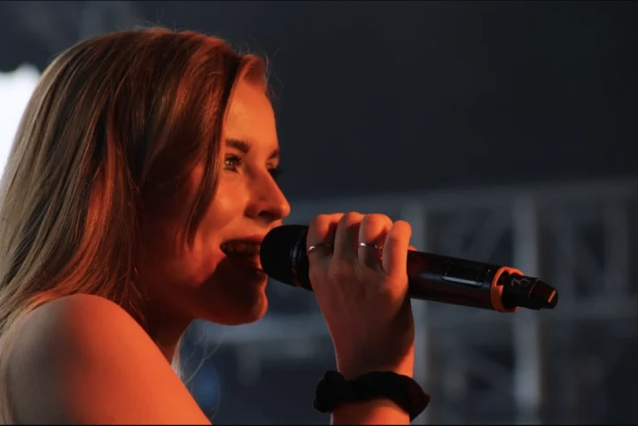 Grace George in profile singing into a handheld microphone, lit in warm orange light against a dark stage.