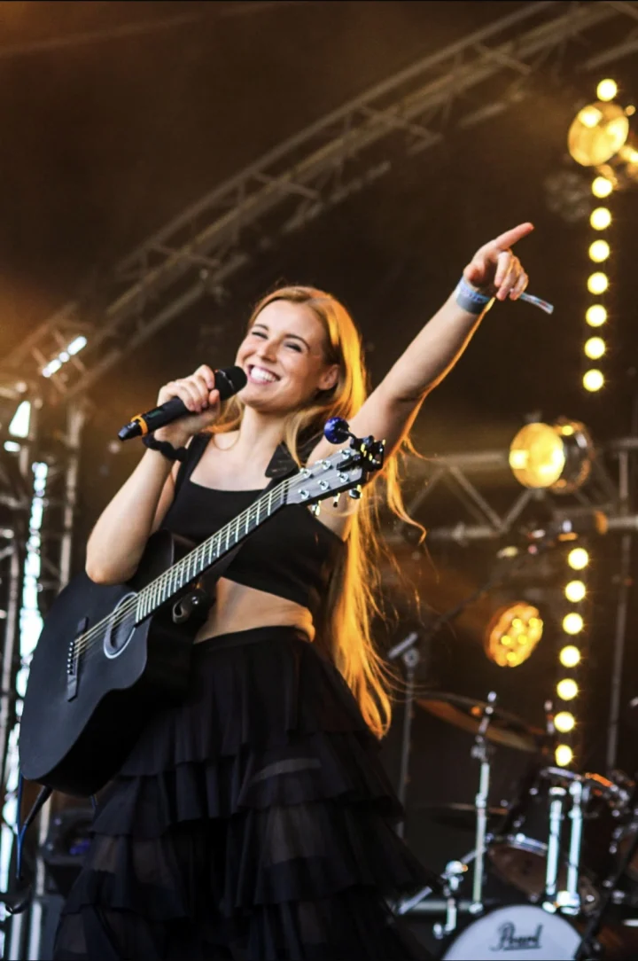 Grace George beaming and pointing toward the crowd, holding a microphone and black guitar on a lit festival stage.