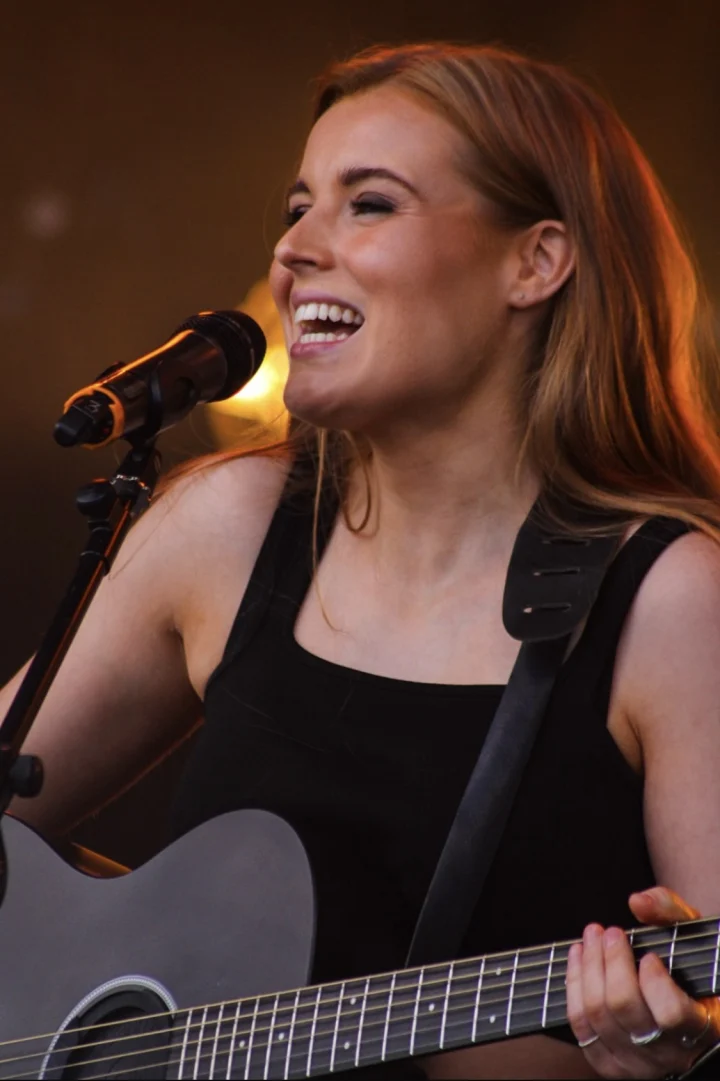 Grace George smiling as she sings into a microphone while playing a black guitar, bathed in warm golden stage light.
