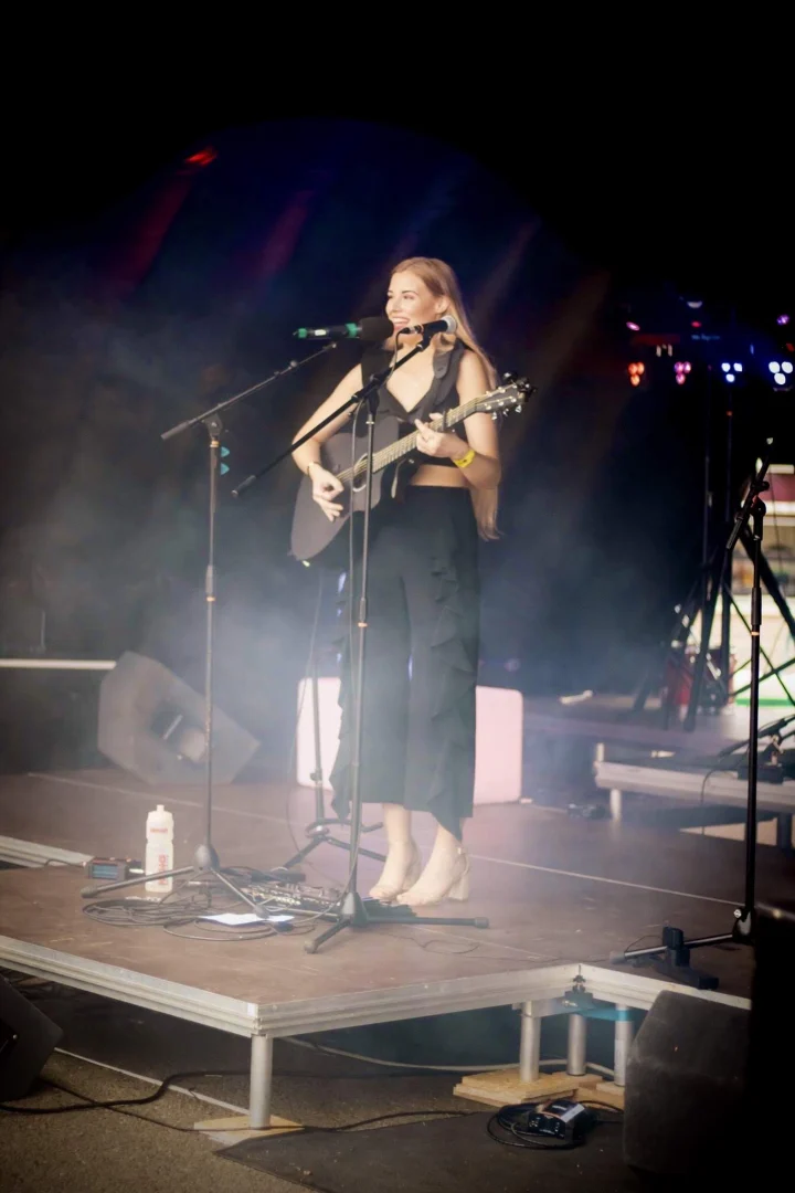 Grace George performing with an acoustic guitar at two microphones on a smoky outdoor stage, in a black ruffled outfit.