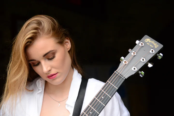 Grace George with eyes closed, looking down beside the headstock of a Martin guitar, wearing a white shirt, dark background.