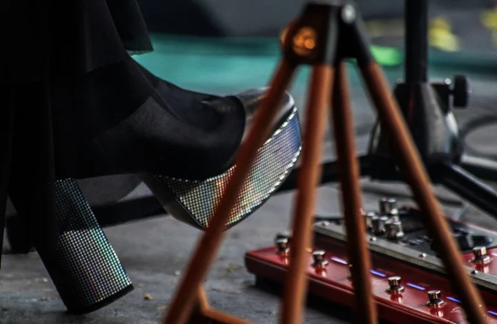 Close-up of a glittery platform boot poised above a red multi-switch foot pedal on stage, behind a tripod mic stand.