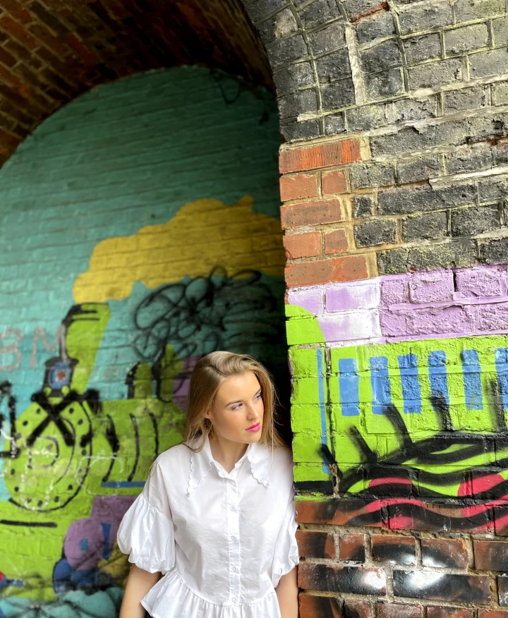 Grace George leaning against a brick arch covered in bright green, yellow, and purple graffiti, wearing a white ruffled blouse.