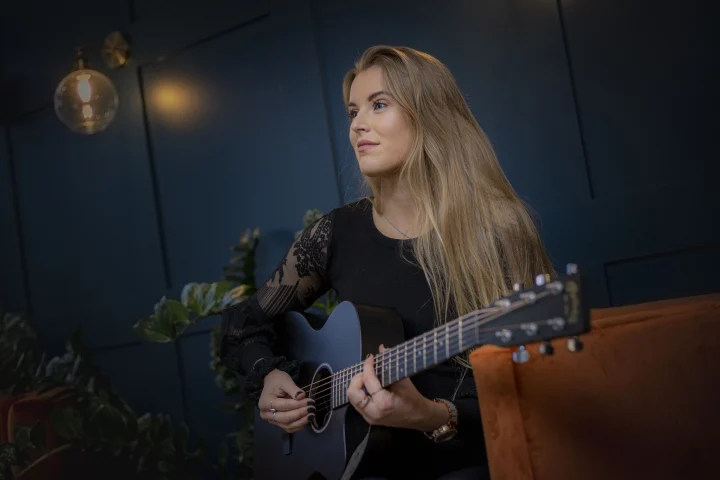 Grace George playing an acoustic guitar in a lounge with a dark teal paneled wall, glowing globe lamp, and plants.