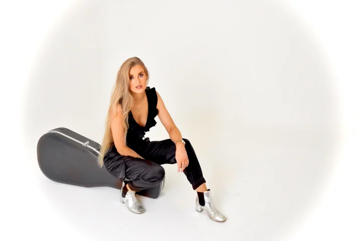 Grace George crouching beside a black guitar case in a white studio, wearing a black jumpsuit and silver ankle boots.