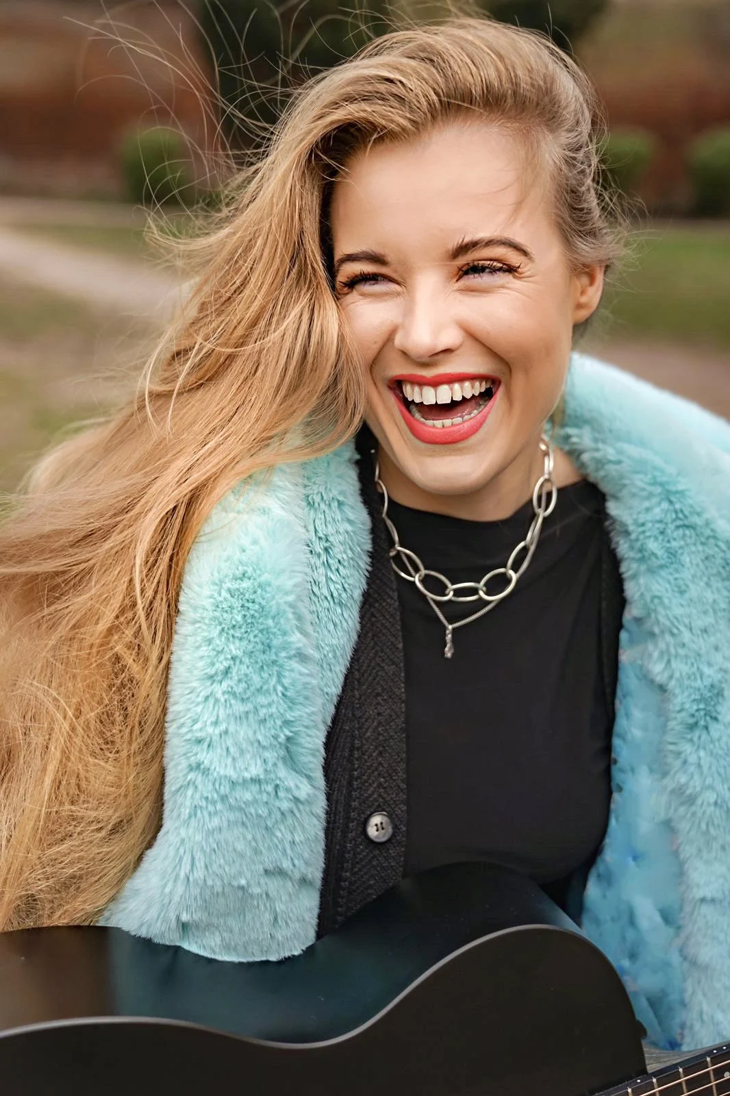Grace George laughing with windswept blonde hair, wearing an aqua faux-fur coat and chain necklace, holding a black guitar outdoors.