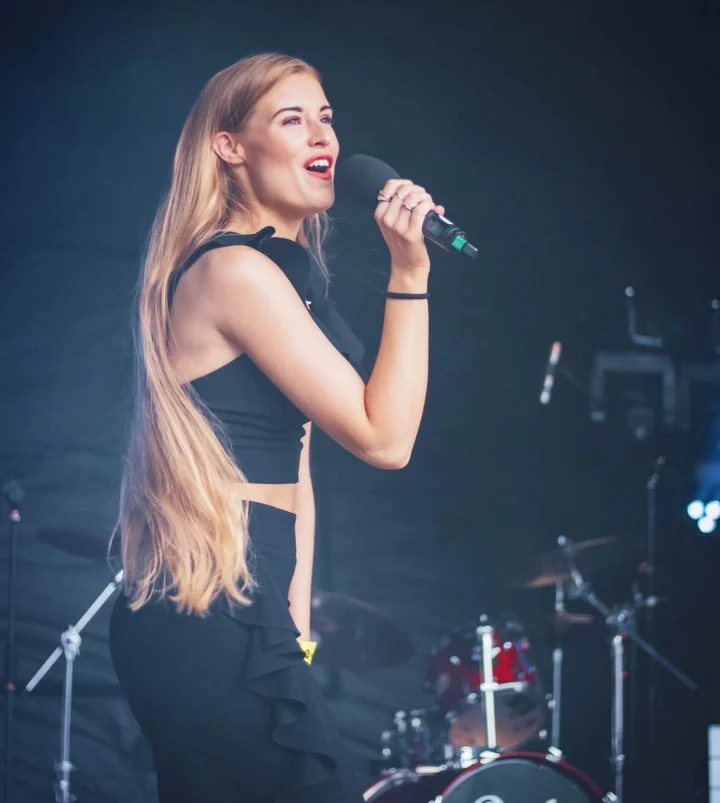 Grace George singing into a handheld microphone on an outdoor stage, wearing a black crop top, drum kit behind her.