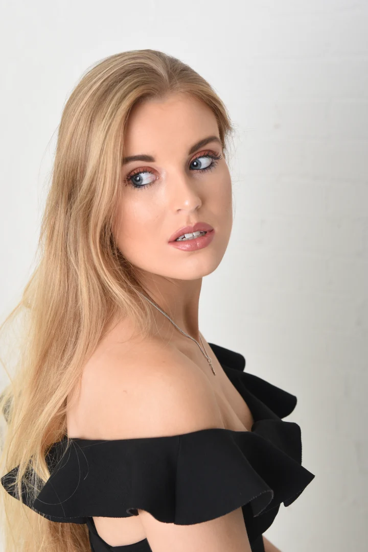 Grace George glancing back over a bare shoulder in a black off-shoulder ruffled top, long blonde hair, on white.