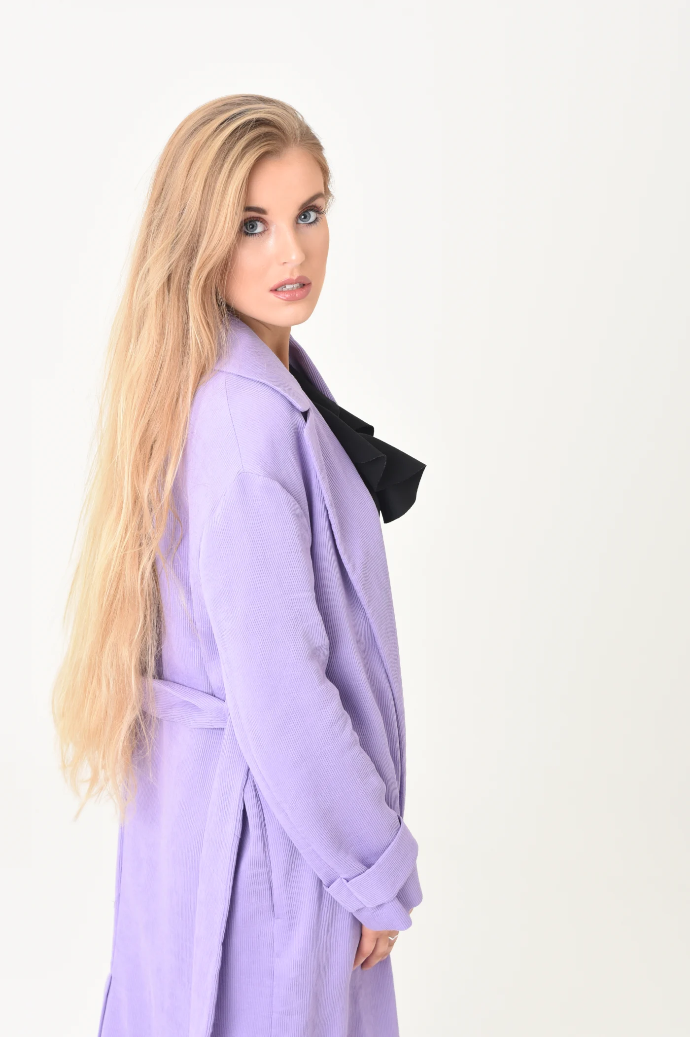 Grace George looking over her shoulder at the camera in a lilac corduroy coat, very long blonde hair, on white.