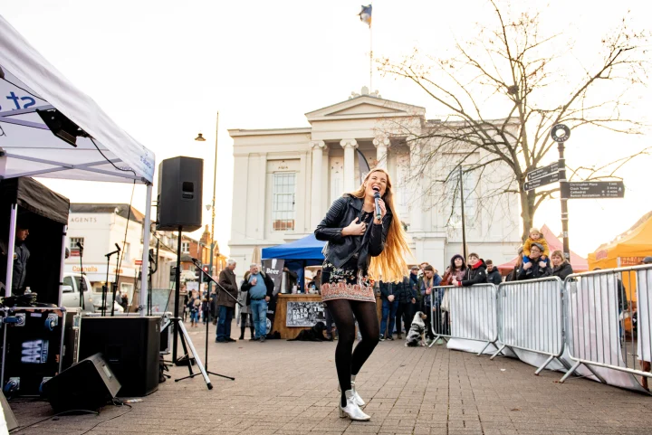 Grace George singing into a microphone on a paved square in a leather jacket, crowd behind barriers, columned building behind.