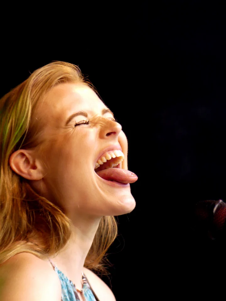 Grace George laughing with eyes squeezed shut and tongue out in a playful close-up against a black background.