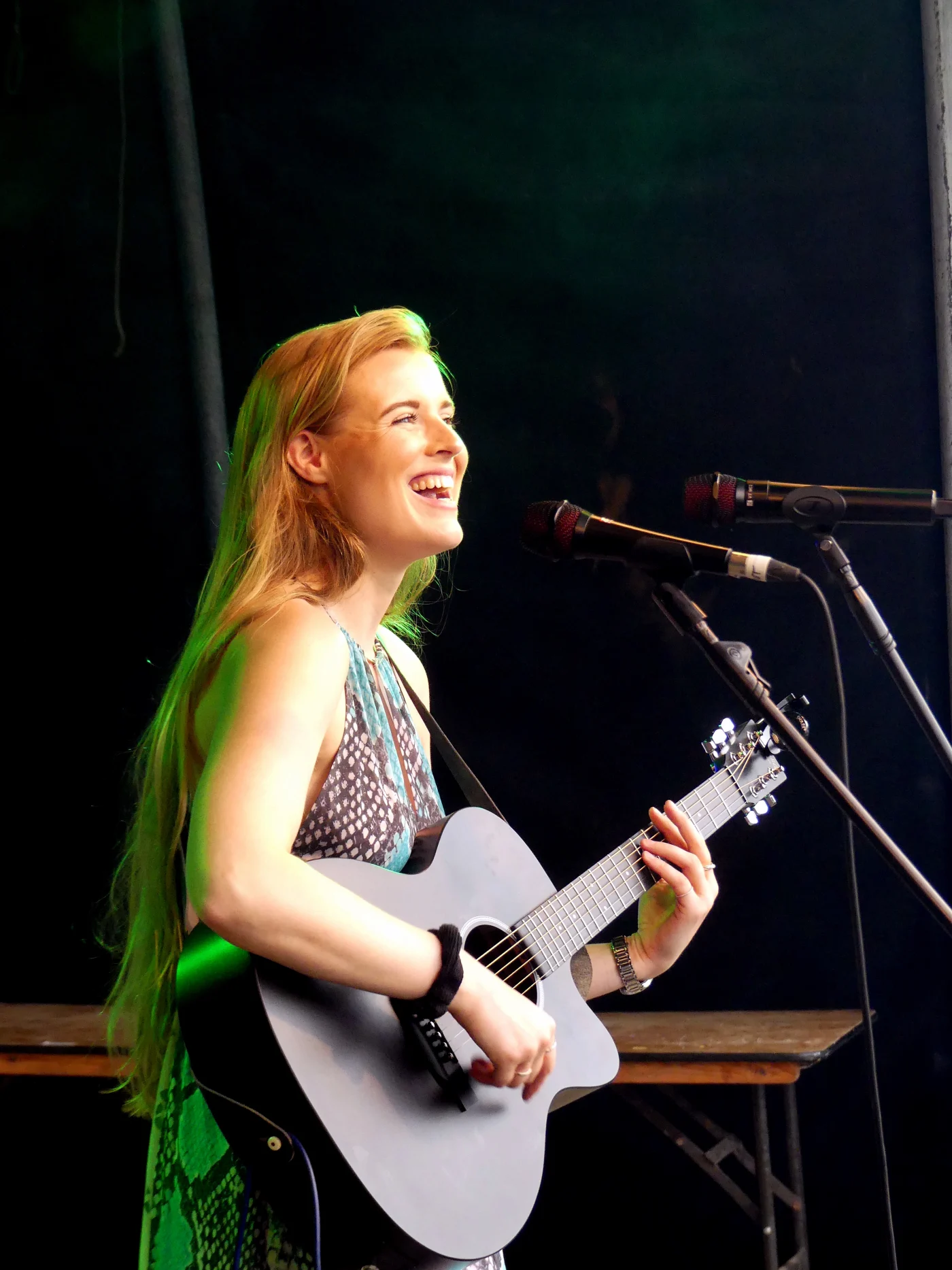Grace George laughing while playing a gray acoustic guitar at two microphones on stage, wearing a patterned halter top.