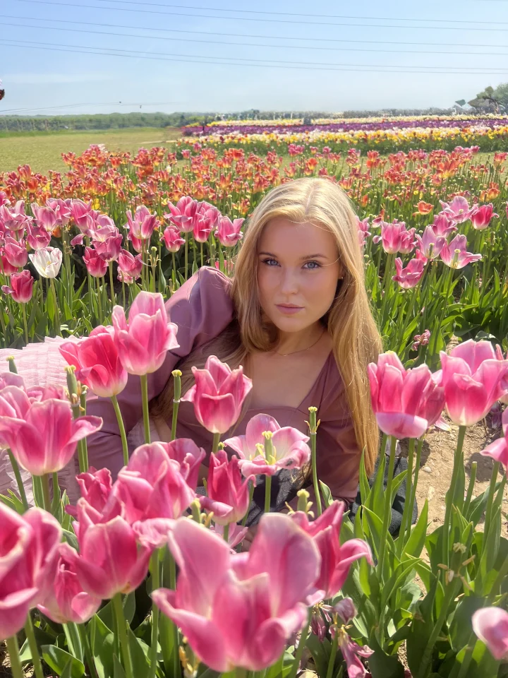 Grace George sitting among pink tulips in a flower field, wearing a mauve blouse and looking at the camera.