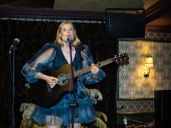 Grace George singing at a microphone while playing guitar, wearing a blue tulle dress with sheer sleeves, art-deco wallpaper behind.