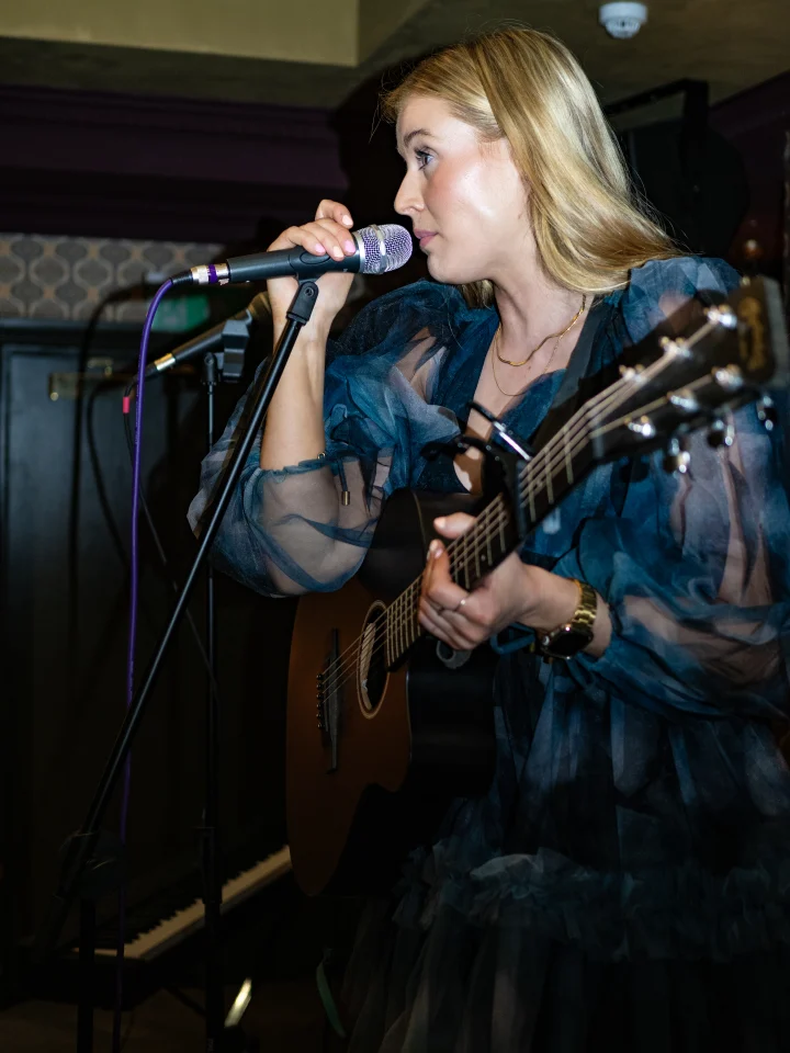 Grace George, seen from the side, singing into a microphone while playing guitar in a blue tulle dress.