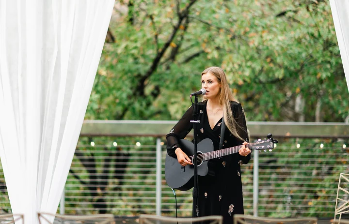 Grace George singing with a black acoustic guitar on an outdoor deck, framed by white drapes and green trees.
