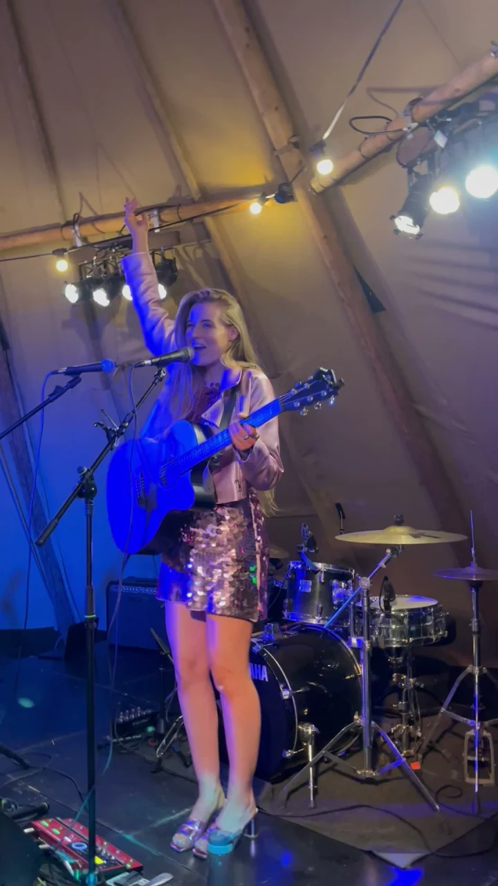 Grace George smiling with one arm raised, singing at a microphone with a blue guitar under a tipi-tent stage rig.
