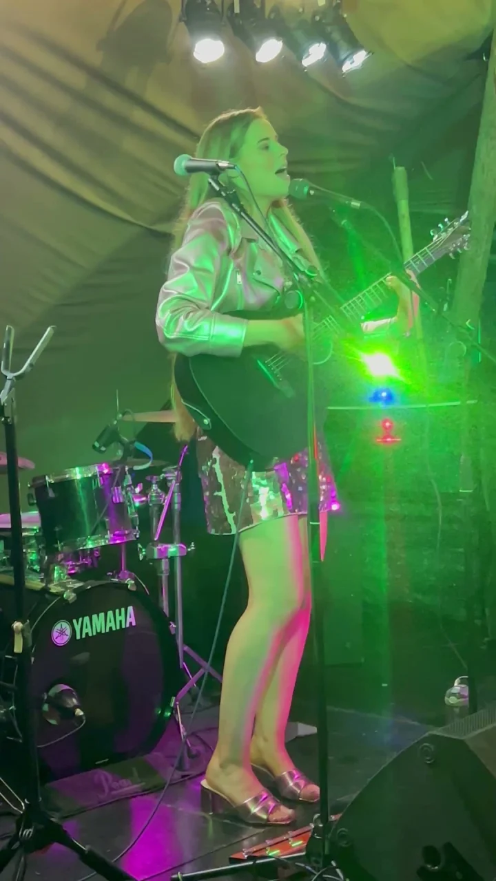 Grace George singing into a microphone with a black guitar under green stage light, a Yamaha drum kit beside her.