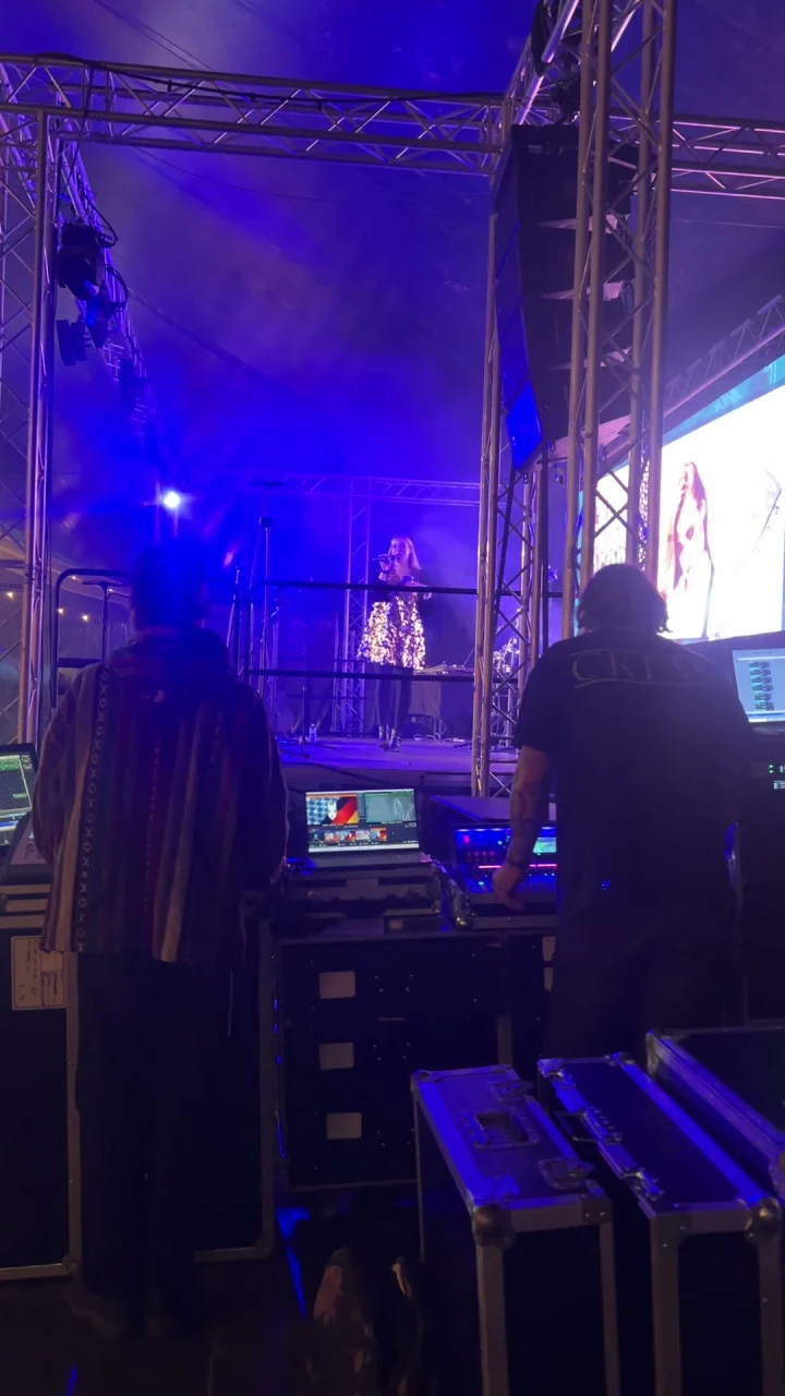 View from behind a sound desk toward a blue-lit festival stage where Grace George performs in a sparkly dress.