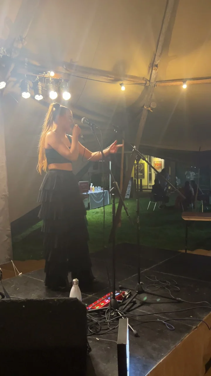 Grace George in a black crop top and tiered skirt, singing into a microphone on a tipi-tent stage.
