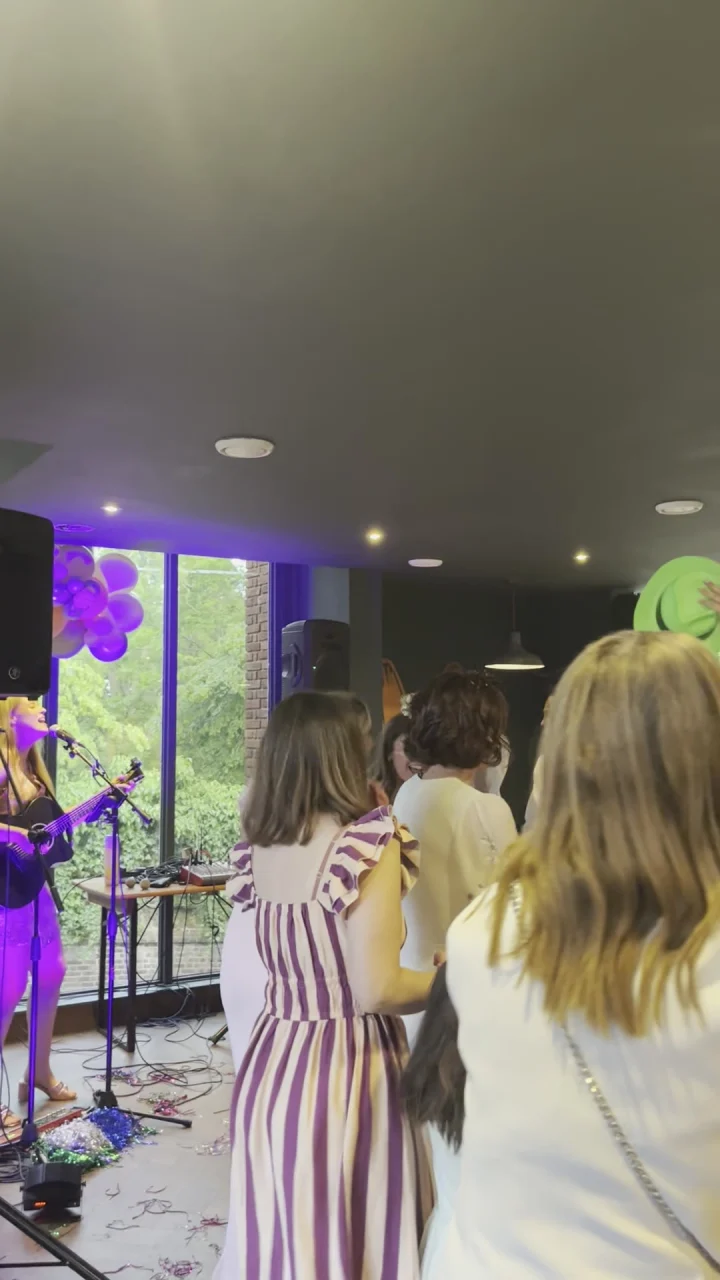 Guests dance in a bright function room with purple balloons while Grace George plays guitar at the left edge.