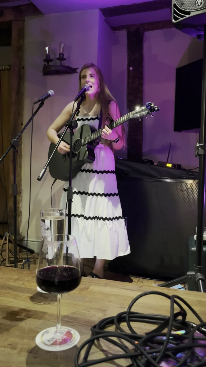 Grace George singing and playing a black guitar in a white dress with black trim, under purple pub lighting.