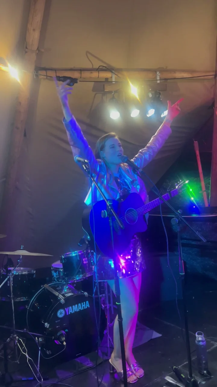 Grace George with both arms raised in triumph, guitar strapped on, singing under colored lights in a tent.