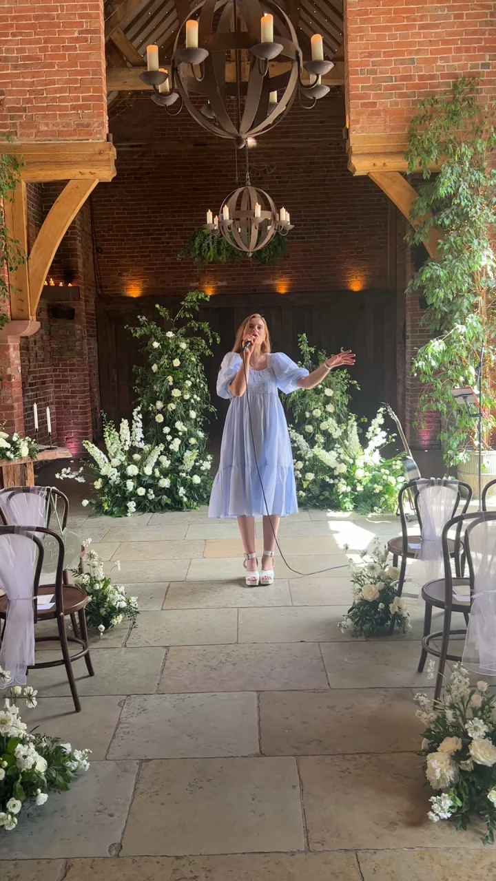 Grace George singing with a handheld microphone in a pale blue dress at the end of a flower-lined barn aisle.