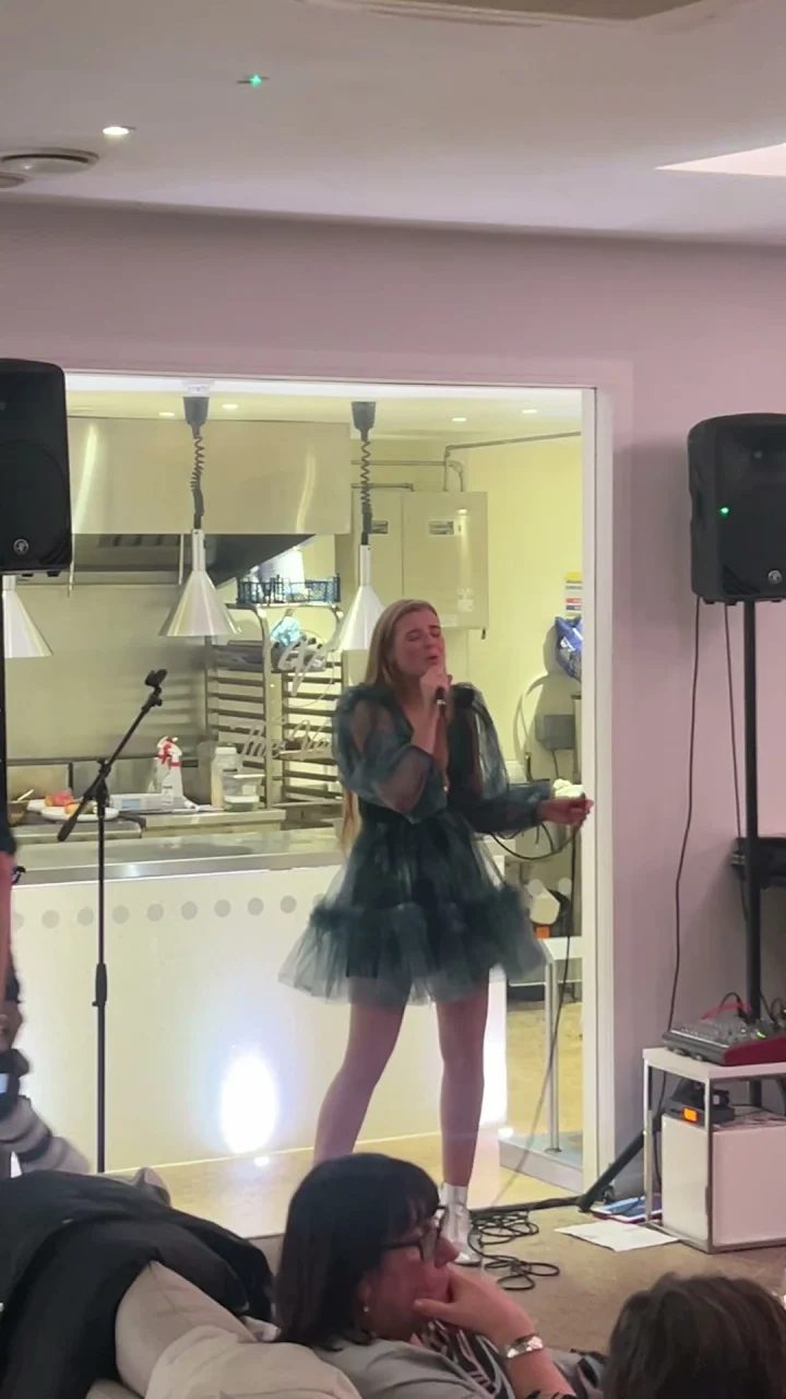 Grace George singing with a handheld microphone in a dark teal tulle dress, in front of seated guests.