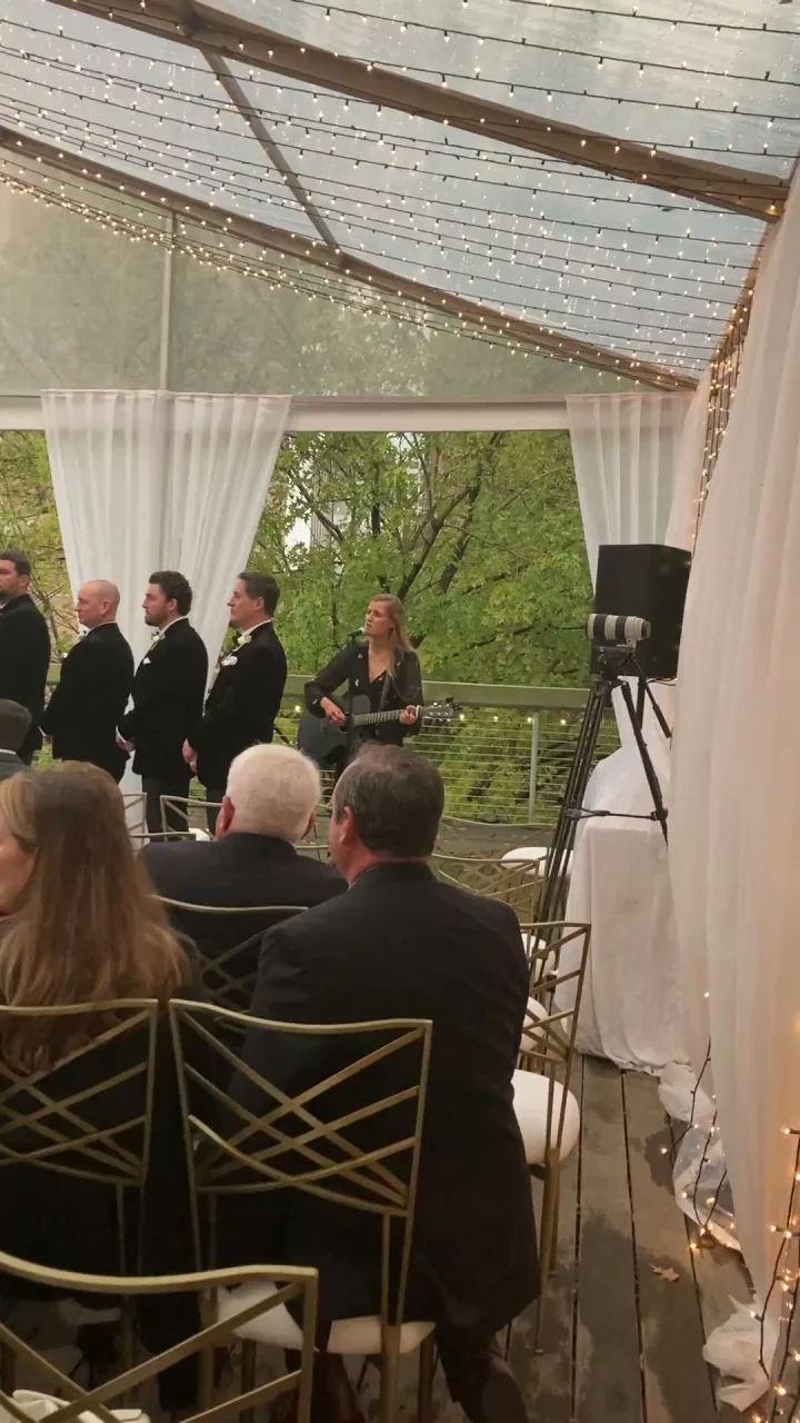 Grace George playing guitar and singing beside groomsmen in tuxedos under a clear tent with fairy lights.