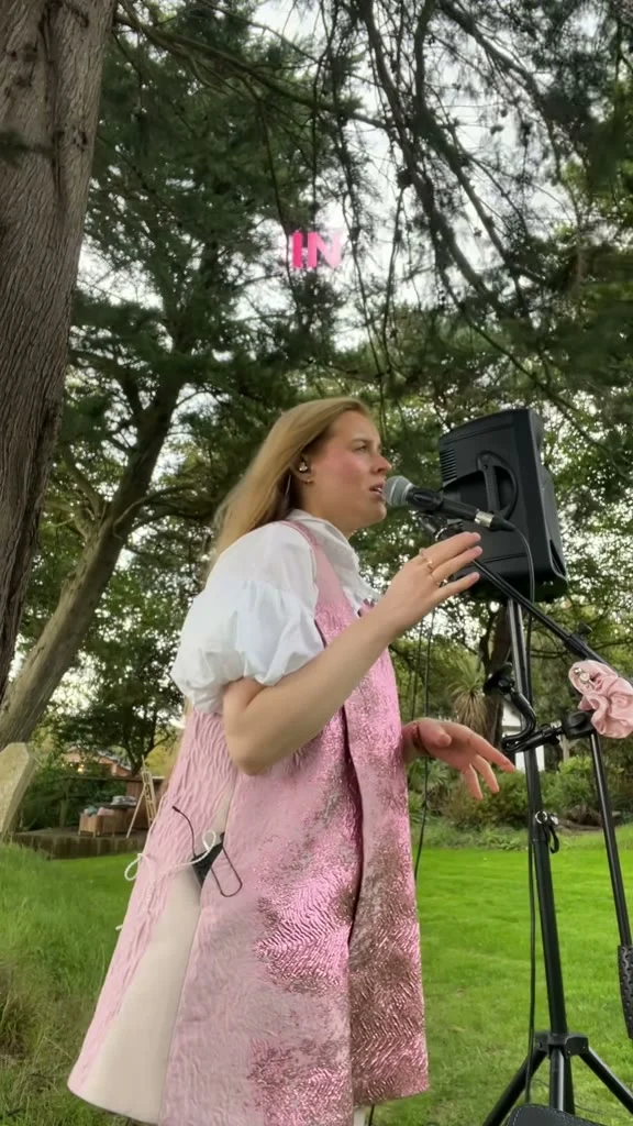 Grace George singing at a microphone in a pink brocade dress with white puff sleeves, in a tree-lined garden.