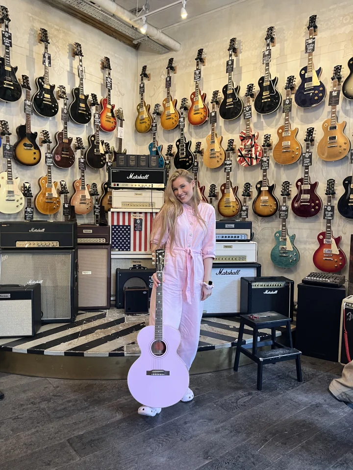 Grace George in a pink outfit with a white guitar, standing in front of a guitar shop wall hung with electric guitars.