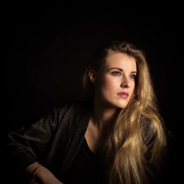 Grace George gazing to the right against a black background, in a dark wrap and a beaded bracelet, long blonde hair falling over one shoulder.