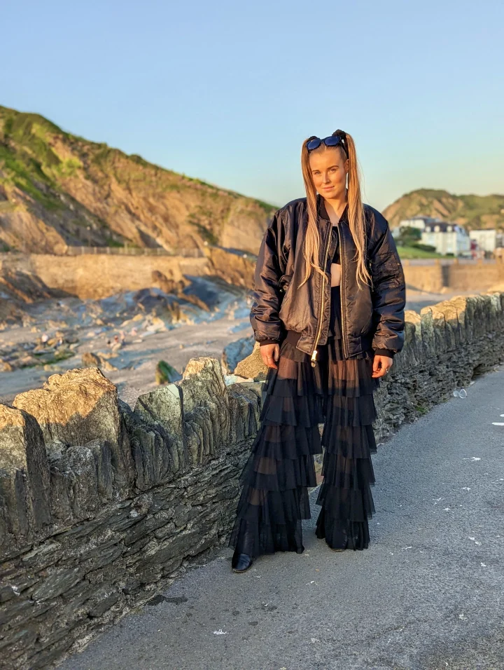 Grace George on a seaside wall in a black jacket and tiered skirt, with cliffs and the sea behind her at sunset.