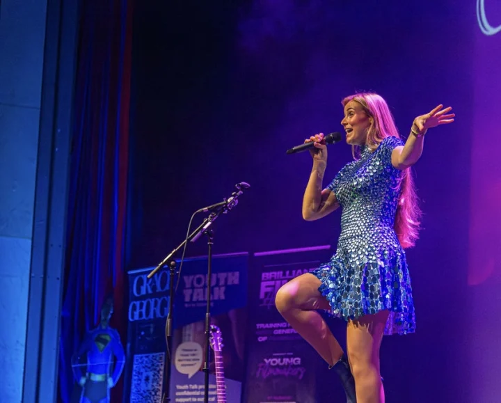 Grace George on a stage lit purple, singing into a handheld microphone in a sequin dress, one arm raised.