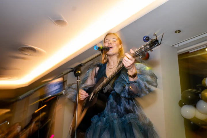 Grace George singing at a microphone in a dark tulle dress, playing guitar in a venue with warm ceiling lights.