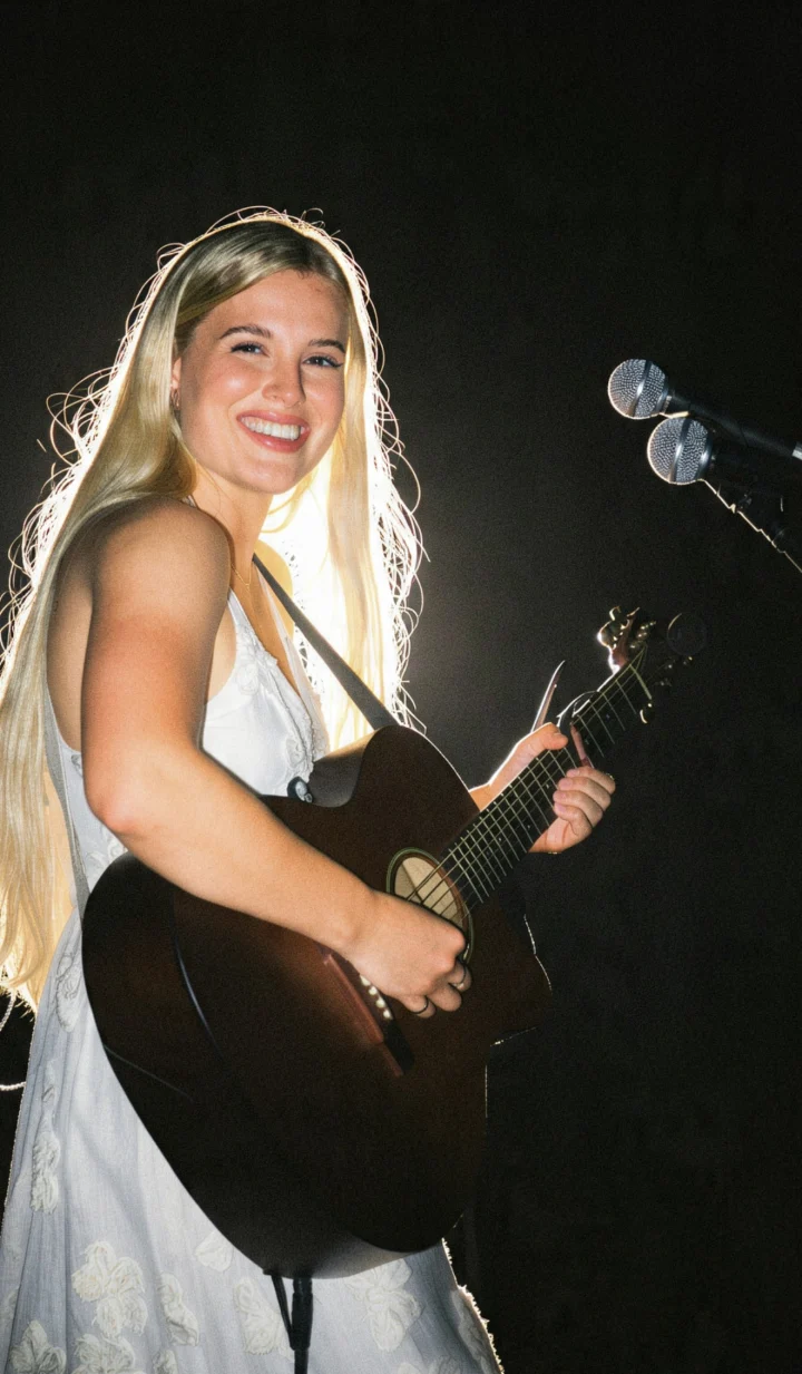 Grace George smiling on a dark stage in a white dress, playing an acoustic guitar beside a microphone.
